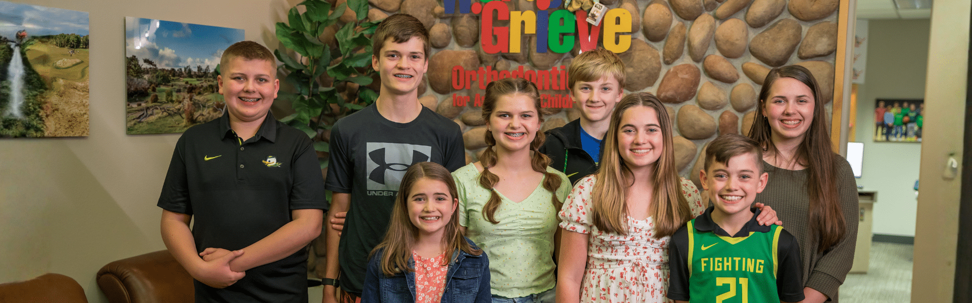 Group of smiling children and teens standing together inside the orthodontic office in front of the practice logo and stone accent wall, Dr. W. Gray Grieve Orthodontics, Eugene, OR