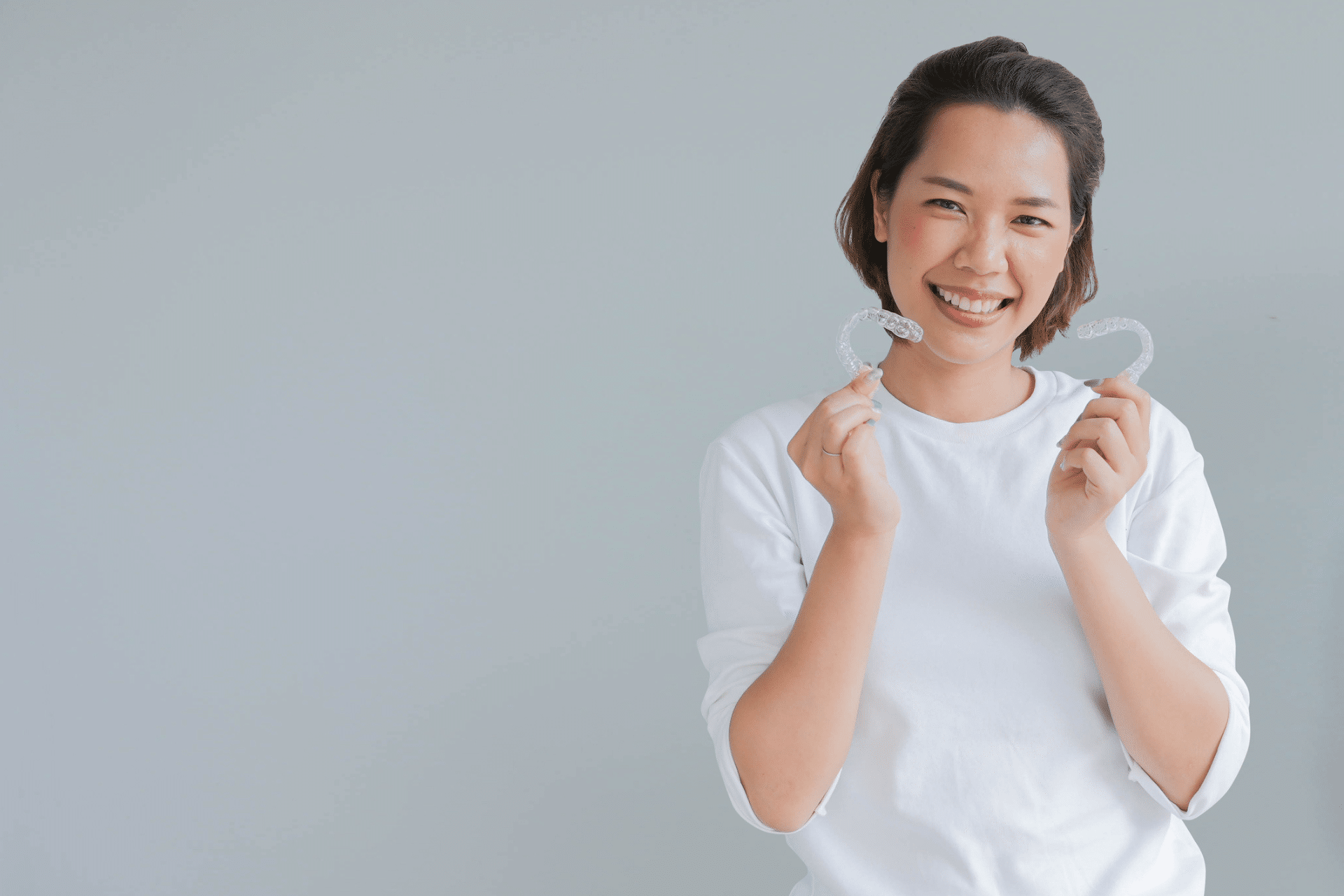 Smiling adult woman holding two clear aligners against a gray background